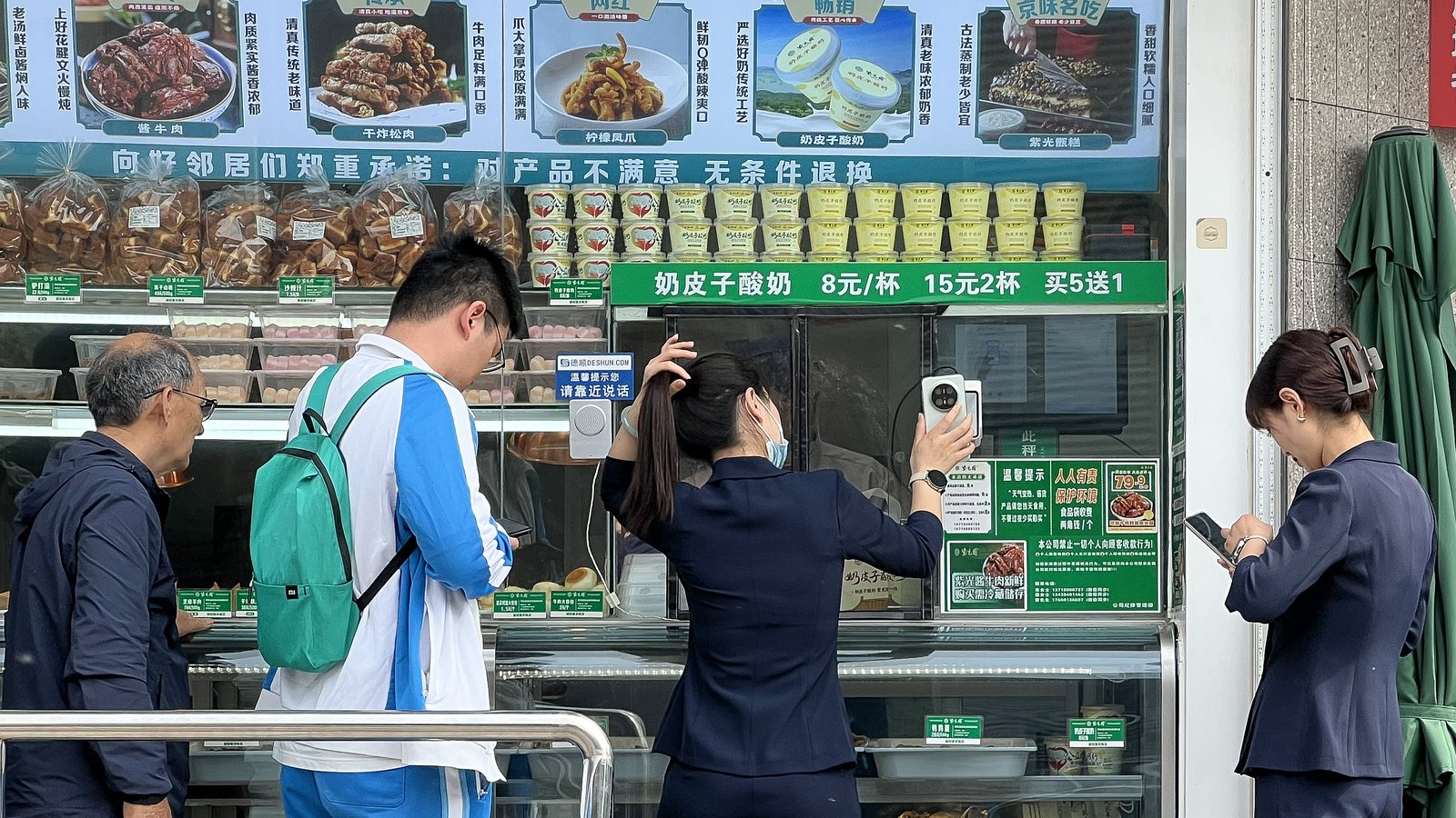 Four customers at the open counter of a Beijing food shop. One woman holds her phone flat against the merchant’s scanner while another waits with her phone already out; behind the glass, chilled trays and a green price board reading 8元 a cup.