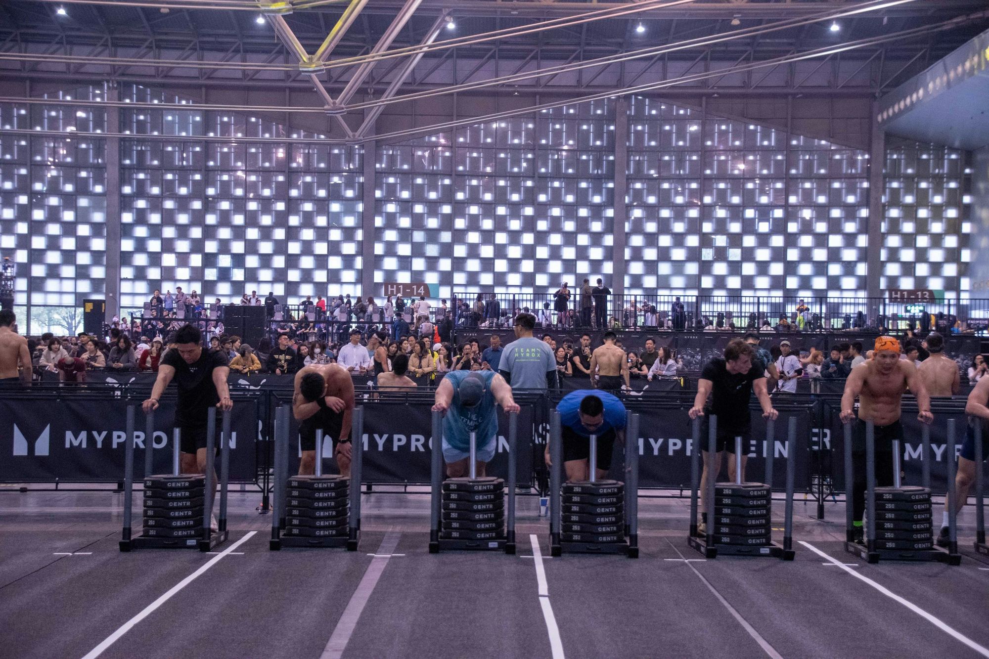 A row of identical weighted sleds lined up across an indoor Hyrox race floor, competitors pushing them down marked lanes in a packed arena