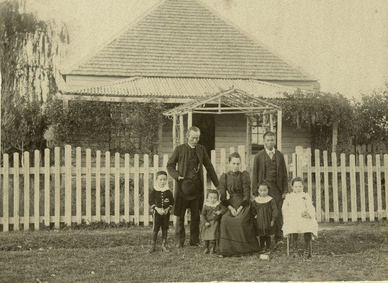 A Chinese family outside their weatherboard home at Inverell: two men in suits, a seated woman in a dark dress and five small children in front of a picket fence.