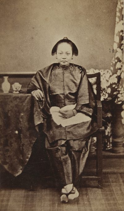 A carte de visite of a young Chinese woman seated beside a draped table, in a dark silk jacket and trousers, holding a string of beads, her bound feet in embroidered shoes.