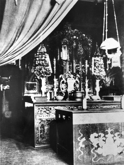 The altar of the Hou Wang temple at Atherton in 1929: carved and gilded woodwork, candlesticks, vases of paper flowers and a hanging lamp, with a painted offering table in front.