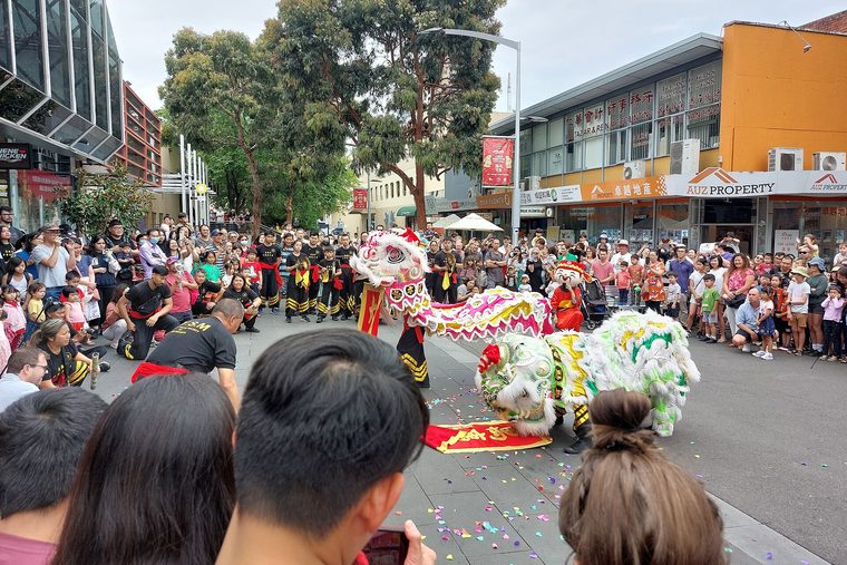 A lion dance for Chinese New Year in Australia, on Bank Street in Box Hill, Melbourne: two lions in front of a crowd and bilingual shopfronts.