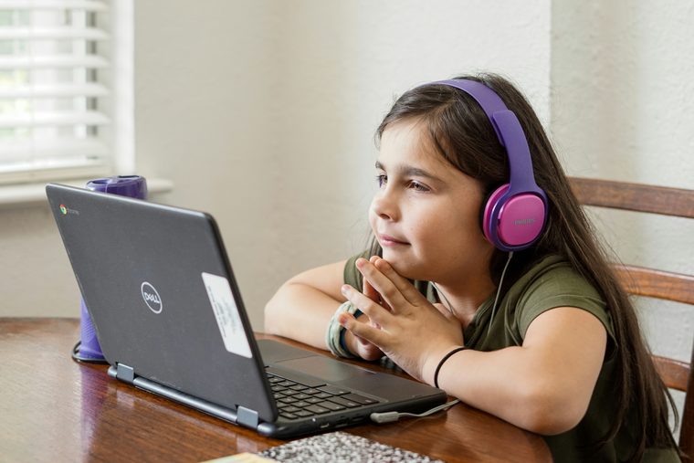 Mandarin for kids: a girl in purple headphones at a kitchen table, chin on her hands, listening to a lesson on her school laptop.