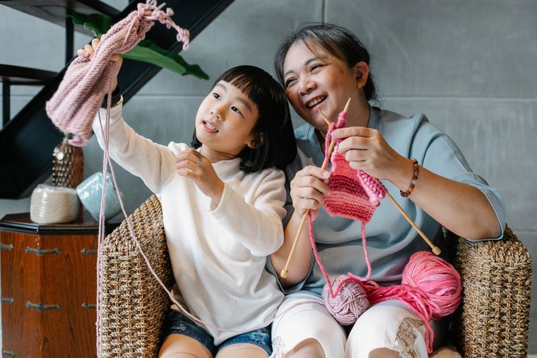 A grandmother and her young granddaughter laughing together over their knitting in a cane armchair.